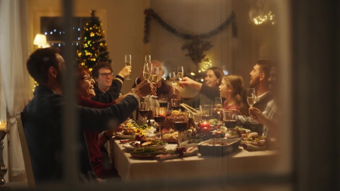 透过雪屋窗户的电影镜头:与父母，孩子和朋友在家里共进圣诞晚餐。多元文化家庭举起香槟酒杯敬酒，庆祝节日