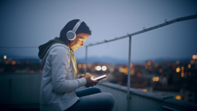女人戴着耳机，晚上在城市的屋顶上听音乐