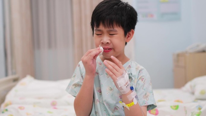 年轻的病人男孩坐在病床上，面对疾病的挑战。生理盐水软管绑在手上为他输送重要液体以帮助他康复。孩子咳嗽