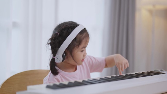 亚洲可爱的女孩微笑着，戴着白色耳机在家里的客厅里学习在线钢琴音乐。隔离期间孩子在家活动的想法。音乐学