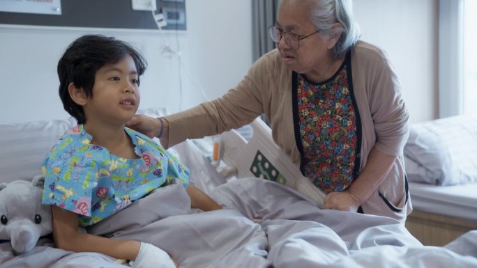 亚洲祖母白天站在病房的病床旁，牵着生病的孙子的手和他说话。