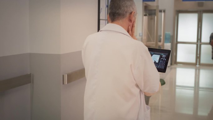 印度医生使用数码平板电脑在医院走廊行走的后视跟踪镜头。资深男性外科医生在手术前检查脑MRI图像的慢动