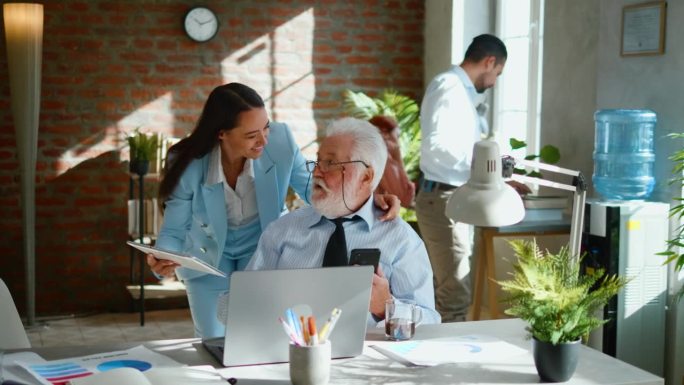 在一间开放式办公室里，一位女秘书拿着平板电脑走向老板的座位，向他征求意见，而老板正在用笔记本电脑工作