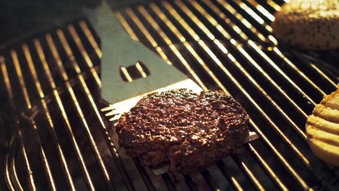 一个人准备汉堡的特写。美味的牛肉饼在火焰烤架上烤，用不锈钢铲翻转肉。电影超级慢动作镜头与速度斜坡效果