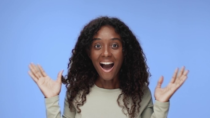 年轻的黑人妇女的肖像看起来兴奋的相机在彩色工作室拍摄。快乐的20岁女孩与哇面部表情孤立独自在蓝色背景