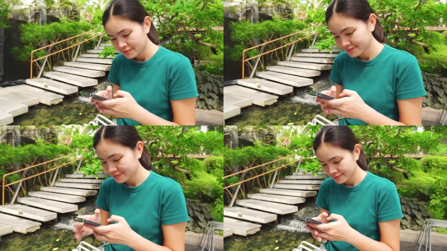 亚洲女人在绿树成荫、流水淙淙的花园里玩手机