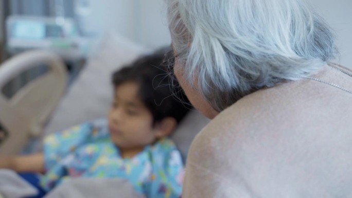 一个生病的小男孩躺在医院的病床上。他的祖母站在他身边，忧心忡忡。