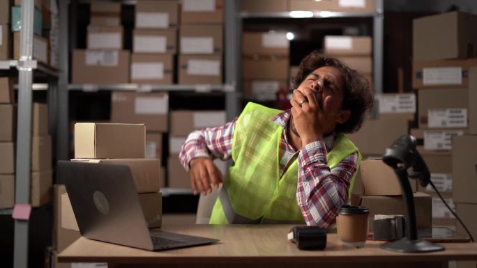 阿拉伯困倦的男工人在纸板箱旁使用笔记本电脑，休息。网上订货，电子商务商品。劳累过度的员工。戏剧性的镜
