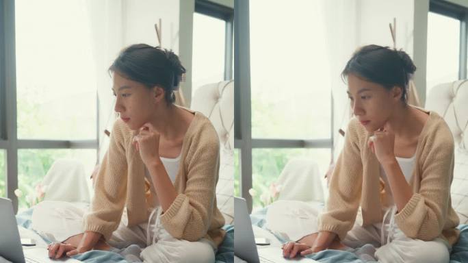 快乐的年轻亚洲女孩，在床上白色奶油开襟羊毛衫，专注电脑，笔记本电脑，满是文书，凌乱的文件，工作的想法