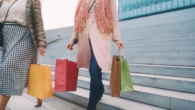两个不知名的女人穿着细高跟鞋拎着购物袋走在城市购物中心前的楼梯上