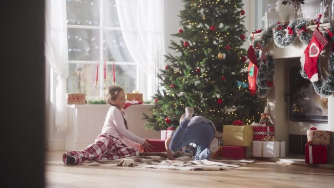 快乐的小弟弟和小妹妹在节日早晨醒来，从圣诞树下收到新玩具的慢镜头。可爱的孩子们迫不及待地去拿礼物