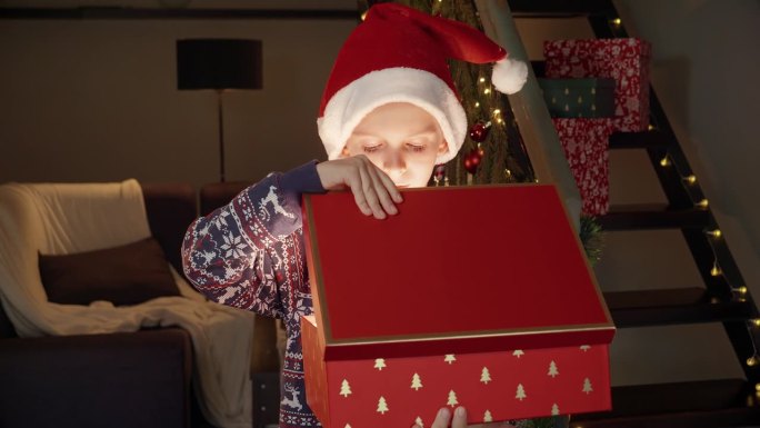 圣诞前夜，戴着圣诞老人帽子的小男孩兴奋地打开了装有礼物的神奇发光的圣诞礼盒。寒假的家庭庆祝活动。