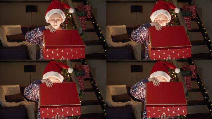 圣诞前夜，戴着圣诞老人帽子的小男孩兴奋地打开了装有礼物的神奇发光的圣诞礼盒。寒假的家庭庆祝活动。
