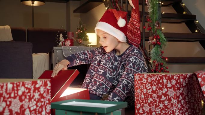 小男孩戴着圣诞老人的帽子，打开圣诞礼盒，兴奋而快乐。寒假的家庭庆祝活动。