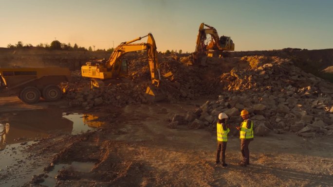 空中无人机拍摄的建筑工地在阳光明媚的晚上:工业挖掘机挖掘岩石，并把它们装上卡车。土木工程师和检查员观