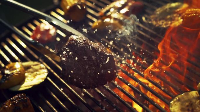 餐厅厨师添加结晶盐片美味的牛排，是准备在火焰烧烤烧烤。创意近距离超级慢动作镜头与速度坡道效果