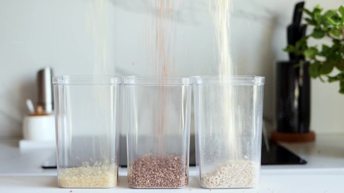 米饭、荞麦和燕麦片被倒入透明的容器中，以现代厨房为背景。健康食品和营养理念。安全、生态、健康产品。生