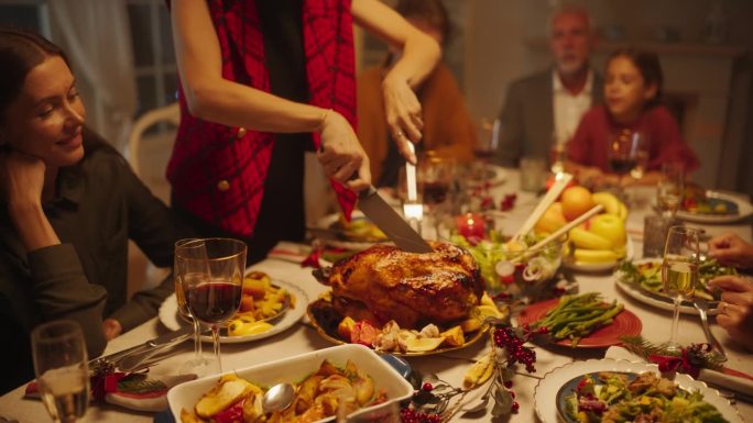 节日感恩节晚餐或圣诞庆祝家庭餐。人们坐在餐桌后面，而一位年轻的女性正在用烤土豆雕刻火鸡大餐。近距离放