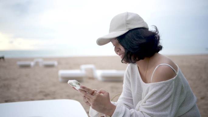 亚洲女游客在海边度假观光。她喜欢用智能手机为她的旅游博客做笔记。放松,享受。他微笑着，看起来很开心。