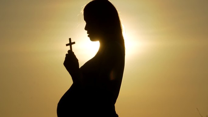 年轻的孕妇在美丽的夕阳下用十字架祈祷