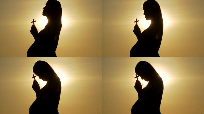 年轻的孕妇在美丽的夕阳下用十字架祈祷