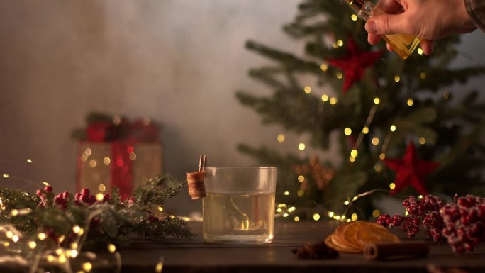 酒保调制圣诞鸡尾酒。在玻璃上洒上金色的喷雾。背景里有花环和礼物的圣诞树。切成片的橘子，肉桂，八角，红