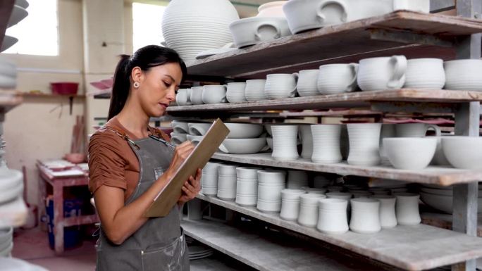 一个漂亮的拉丁美洲员工在一家陶瓷厂工作，在珐琅加工前清点物品，在剪贴板上写下数量