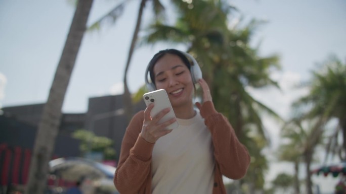 年轻的亚洲女子戴着无线耳机，拿着智能手机在沙滩上跳舞