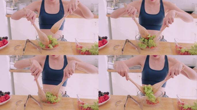 一个穿着运动服的亚洲女人在厨房里快乐地扔着蔬菜沙拉，散发着幸福的光芒。她崇尚健康的生活方式，通过令人
