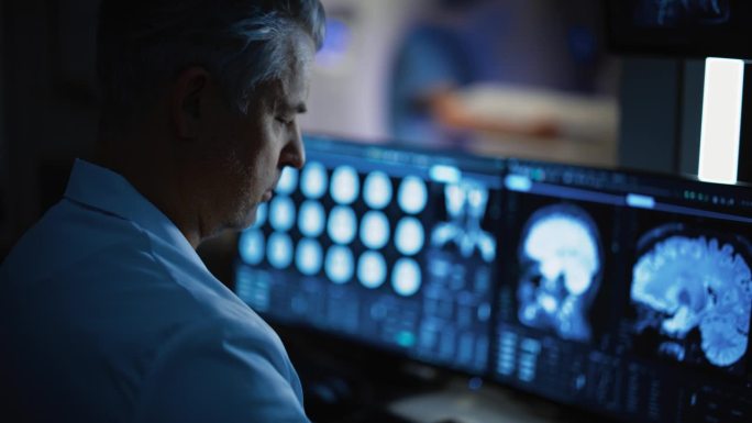 专业医生的特写，看着电脑显示器上的核磁共振或CT扫描图像，寻找脑肿瘤或癌症的迹象，试图诊断疾病