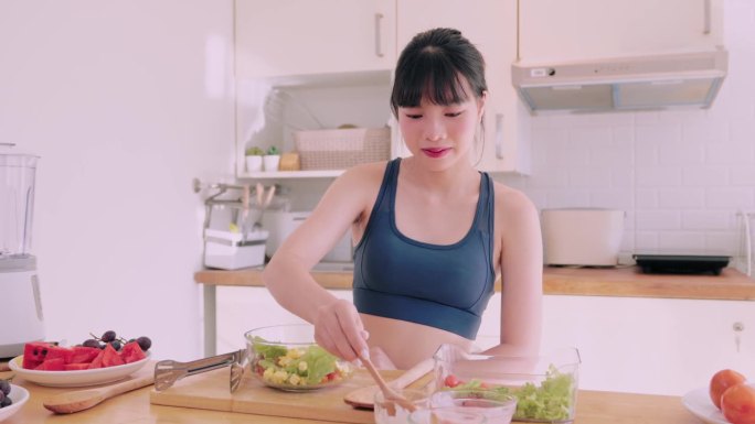 一个穿着运动服的亚洲女人在厨房里快乐地扔着蔬菜沙拉，散发着幸福的光芒。她崇尚健康的生活方式，通过令人