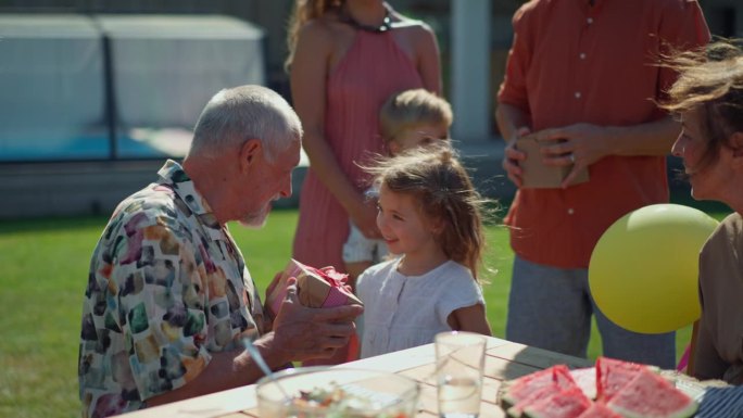 快乐的小女孩在夏季花园的一代家庭生日聚会上给她的老祖父送生日礼物。