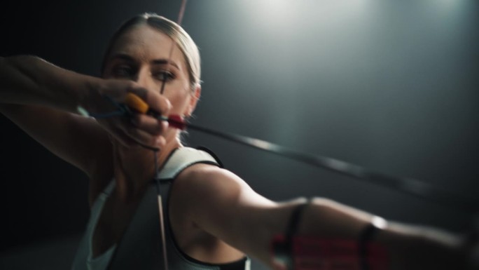 一名女射手手持现代复合弓瞄准目标的电影肖像。年轻运动员在体育场比赛或在黑暗的广告工作室画箭