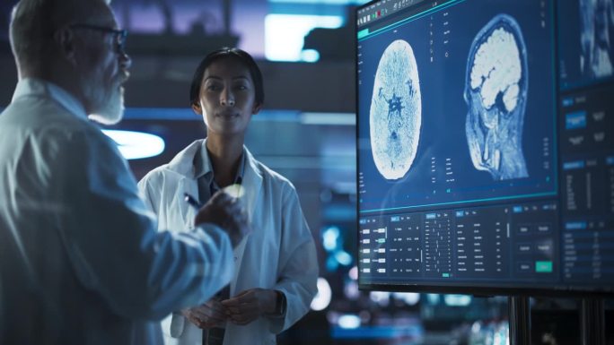 神经科学实验室会议:亚洲女科学家和男外科医生讨论电视显示的核磁共振脑部扫描。研究脑损伤后康复药物的专