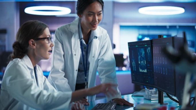 亚洲和高加索女医生在医学研究中心的谈话和使用台式电脑分析脑核磁共振扫描。外科医生和神经科学家讨论治疗