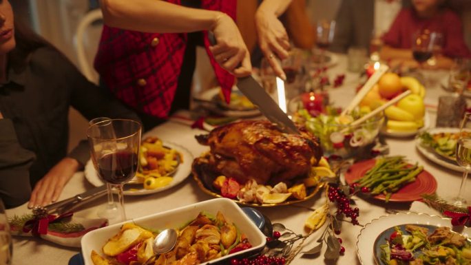 快乐的客人坐在餐桌上享用美味的饭菜和节日的装饰。年轻的女性正在雕刻一只火鸡，准备在家里的家庭晚餐上庆
