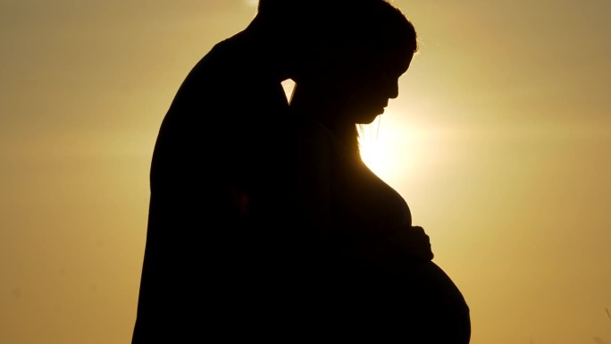 夕阳剪影中，丈夫亲吻妻子怀孕的肚子。