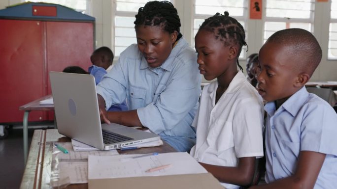 特写镜头。在非洲的一间教室里，非洲黑人女教师坐在桌前，用笔记本电脑帮助非洲孩子完成任务