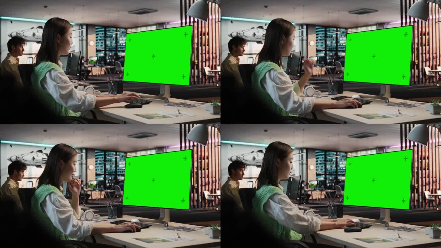 富有创造力的亚洲女性坐在她的办公桌前使用桌面电脑与绿屏Chromakey模型。女关卡设计师在游戏设计