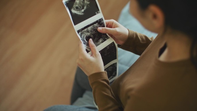 年轻的孕妇在家里的卧室里看着她未出生的孩子的超声波扫描