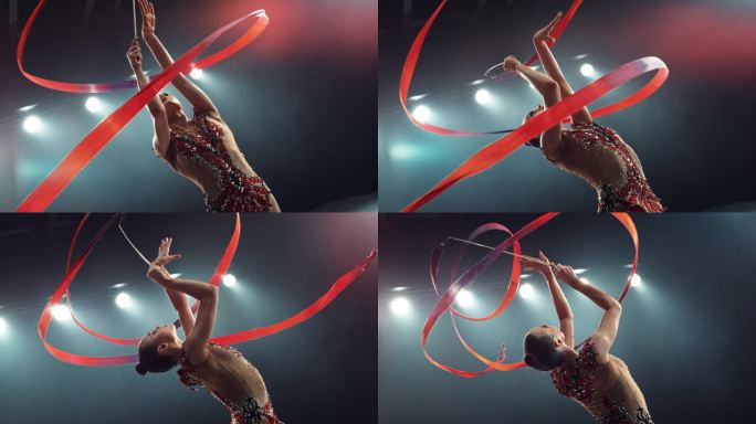 一位年轻美丽的艺术体操运动员系着红丝带表演舞蹈的肖像。年轻女子在黑暗的舞台上穿着醒目的制服纺着丝带。