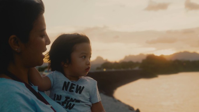亚洲母亲抱着儿子在拉查帕哈大坝看日落。素叻他尼。