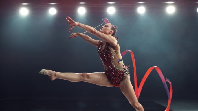 一个现代艺术体操表演的超级慢动作镜头在一个大舞台上的灯光。美丽的女运动员在用红丝带创造形状的同时做出