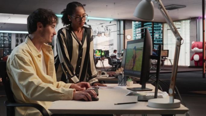 白人男性游戏设计师使用桌面电脑和3D建模软件，并在游戏开发工作室办公室与黑人女性项目经理交谈。不同的
