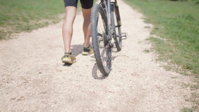 一名男性山地自行车手全副武装地推着他的山地自行车在一块绿地附近的鹅卵石土路上坡。在风景优美的草地上探