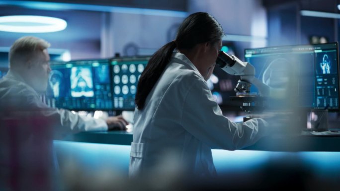亚洲女科学家在生物技术研究中心用显微镜分析培养皿样品。不同的男性同事用台式电脑对大脑进行核磁共振扫描