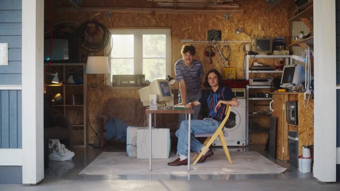 两个白人男性创业公司创始人在复古的车库里使用旧台式电脑，看着相机微笑。程序员和用户体验设计师在90年