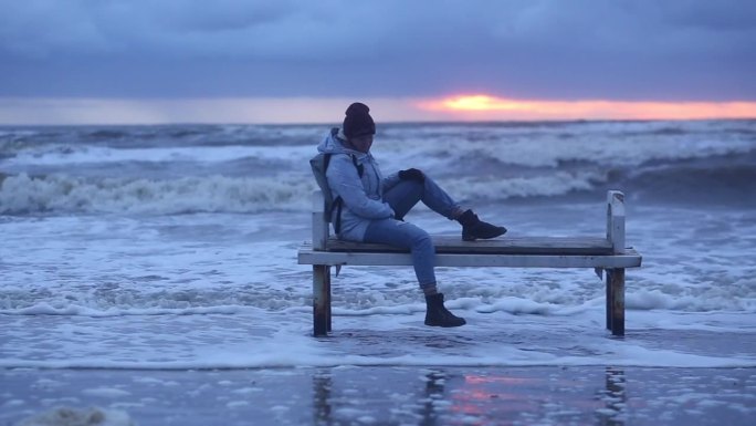 一个孤独的女孩在冬天的暴风雨中坐在海边的长凳上