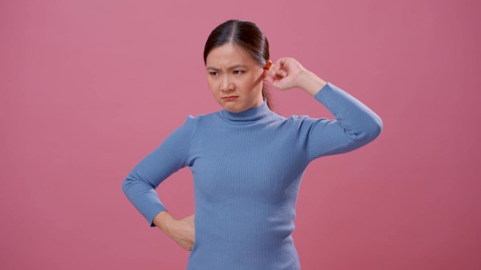 亚洲女人耳朵发痒，把一个手指放进耳朵里，孤立地站在粉红色的背景上。