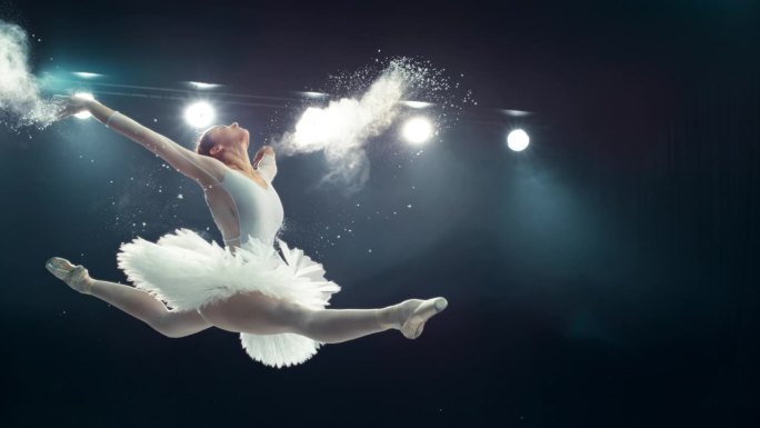 灯光大舞台上美丽的芭蕾舞表演。年轻的女芭蕾舞演员在白色的裙子做一个完美的劈叉跳跃和释放粉末。超级慢动